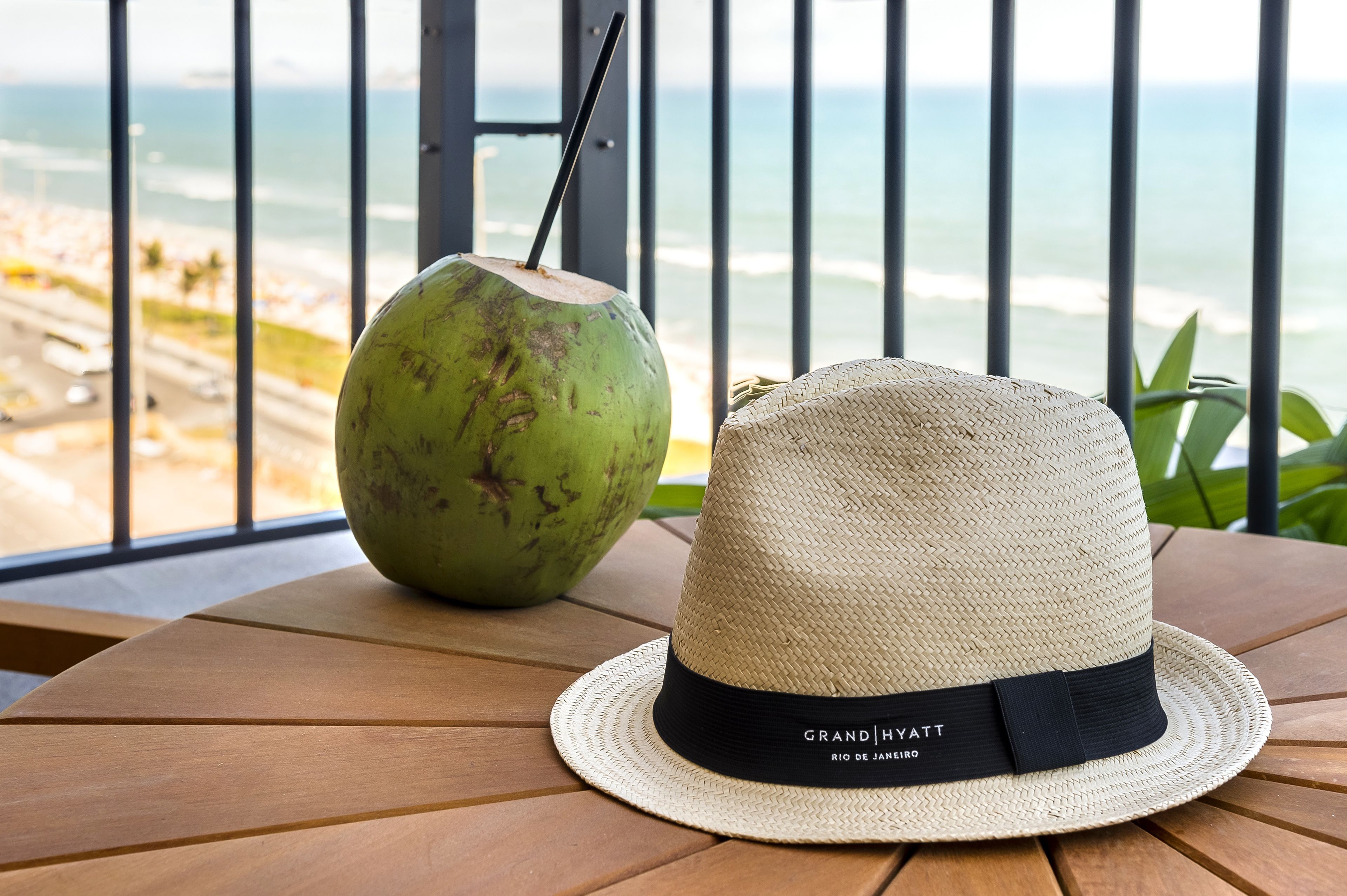 A straw hat and a coconut drink on a wooden table with a view of the beach and ocean.