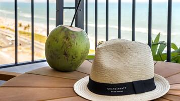 Um chapéu de palha e uma bebida de coco sobre uma mesa de madeira, com vista para a praia e o oceano.