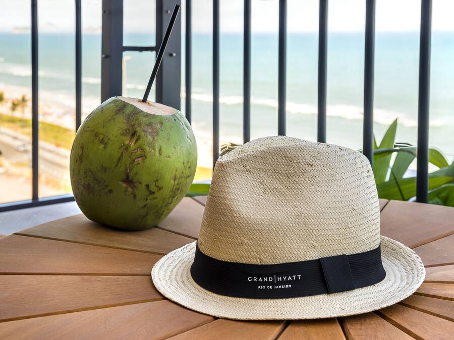 A straw hat and a coconut drink on a wooden table with a view of the beach and ocean.