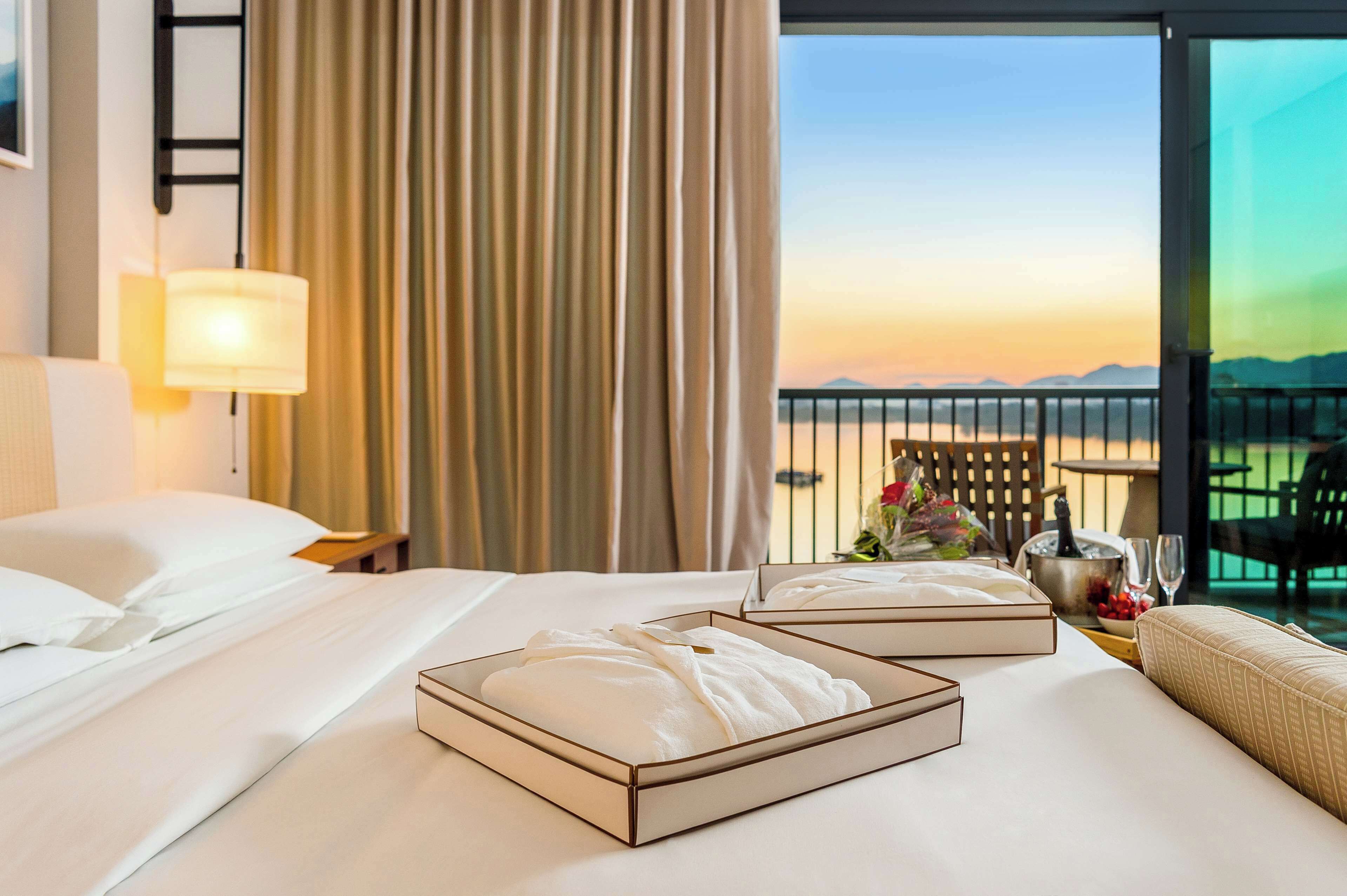 Habitación de hotel con cama, dos almohadas, una mesita de noche con lámpara, vista al atardecer y un balcón con mesa y sillas.