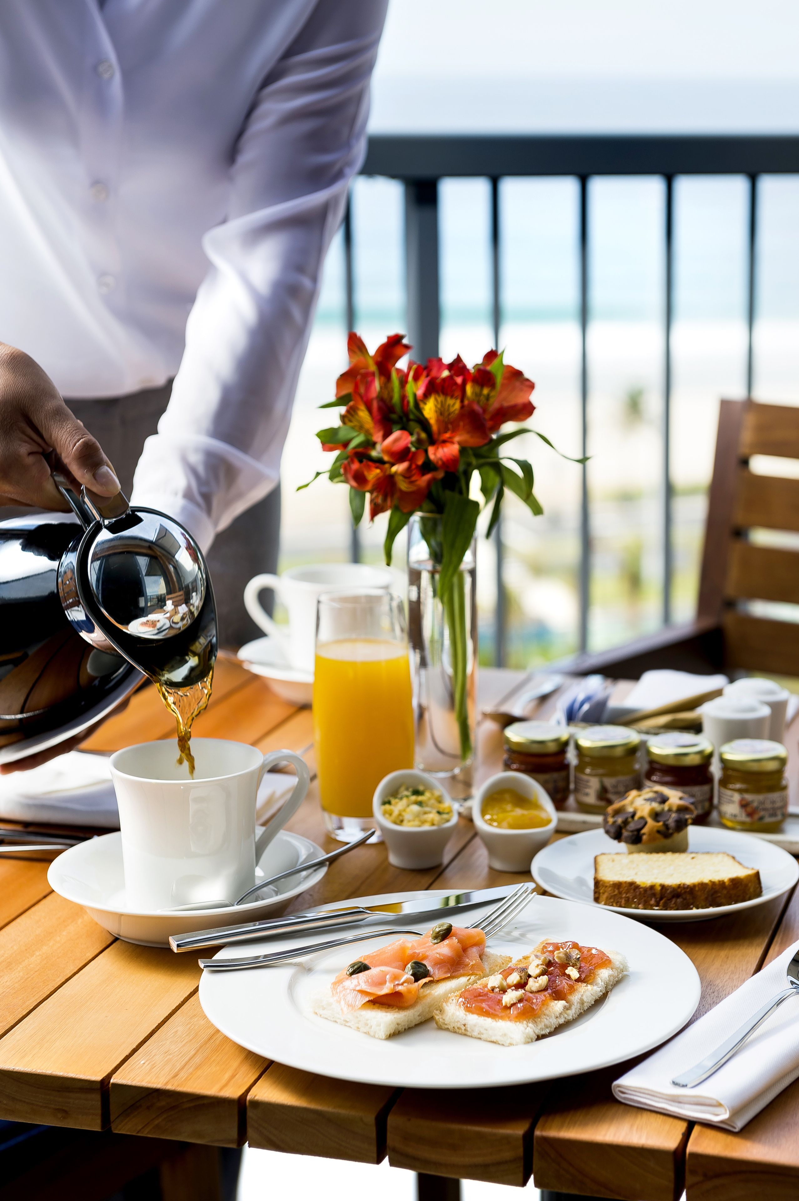 Una colazione con una persona che versa il caffè in una tazza, un vaso di fiori e diversi cibi da colazione su un tavolo di legno.