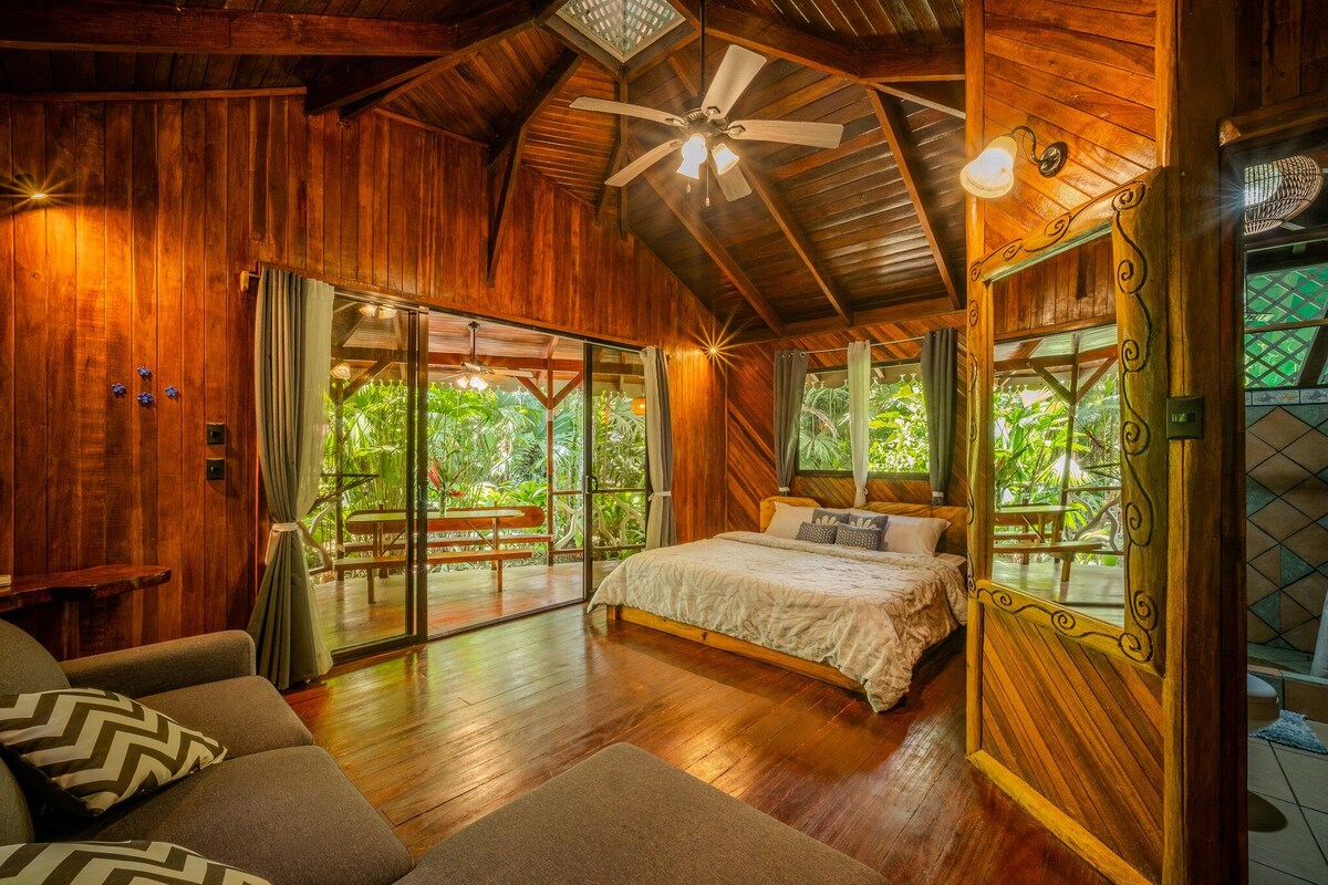 Una habitación de madera con una cama, un ventilador de techo y vistas al verdor exterior.