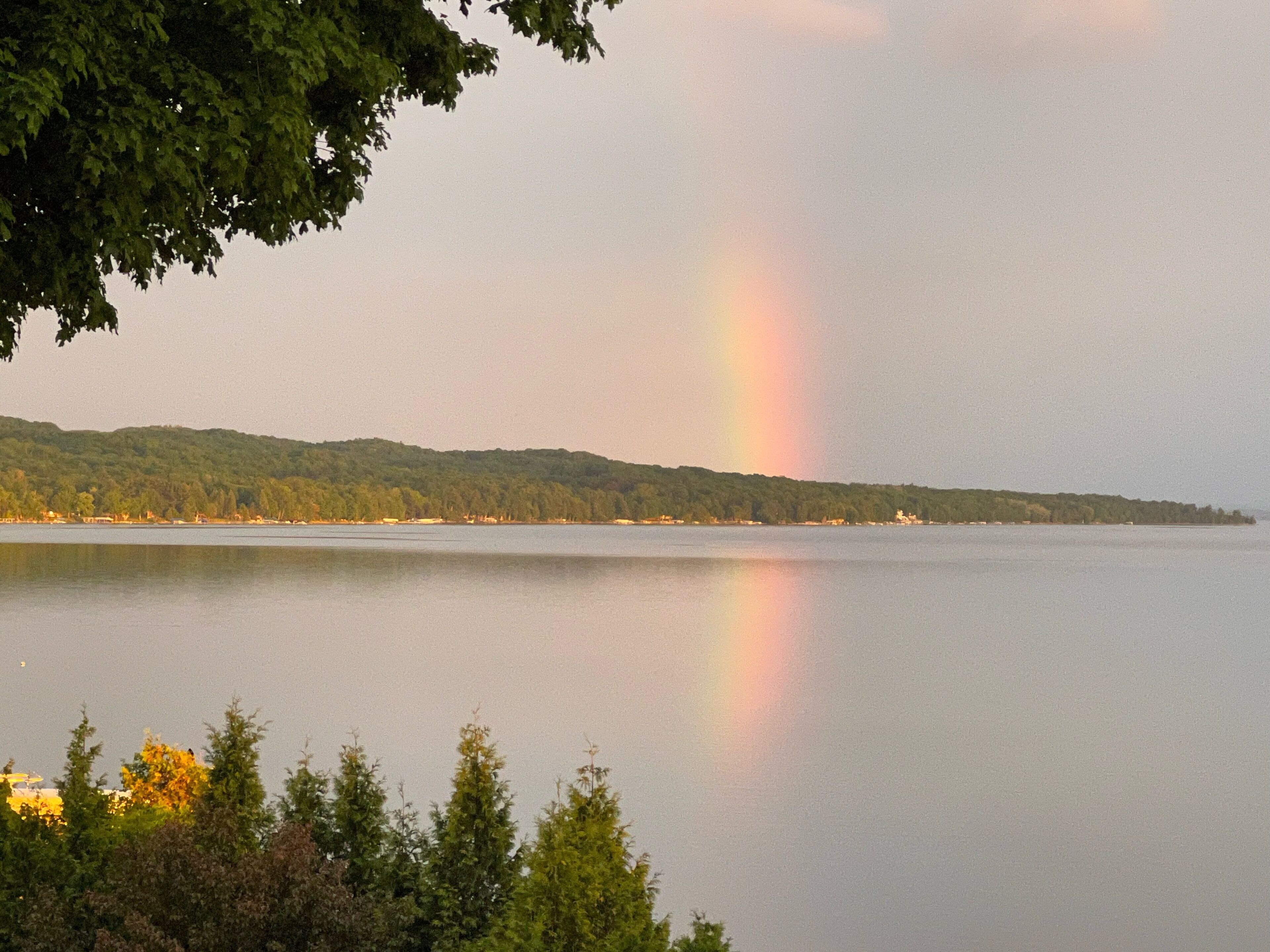 A calm lake with a rainbow in the distance, surrounded by trees and a hilly landscape.