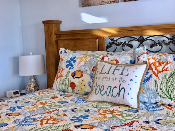 A bed with a colorful floral and sea-themed bedspread, a pillow with the text "LIFE is good at the beach," and a wooden headboard. - Torch Lake Bed & Breakfast (Central Lake)
