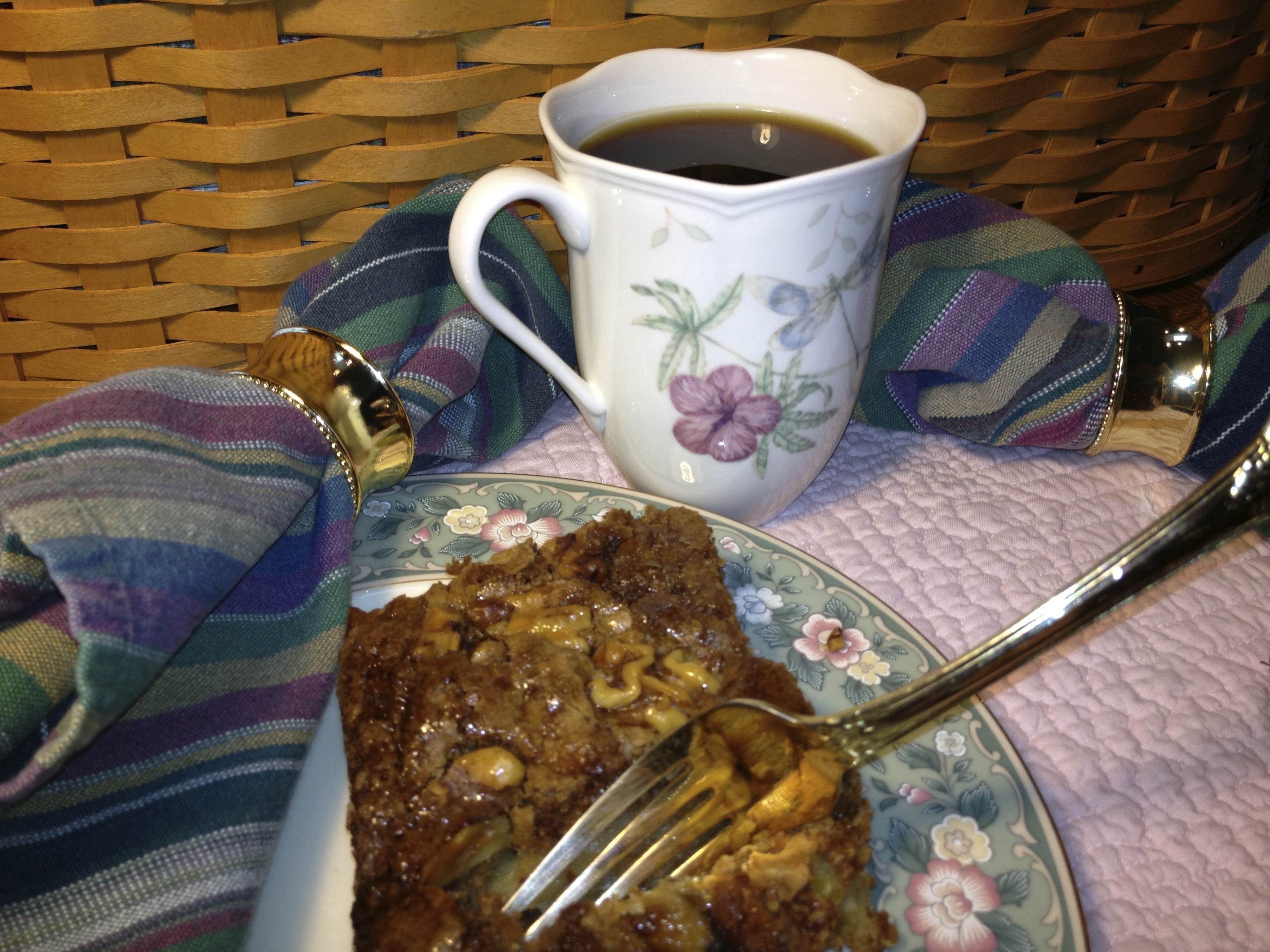 A slice of cake with nuts on a plate, a fork, a cup of coffee, and a striped cloth.