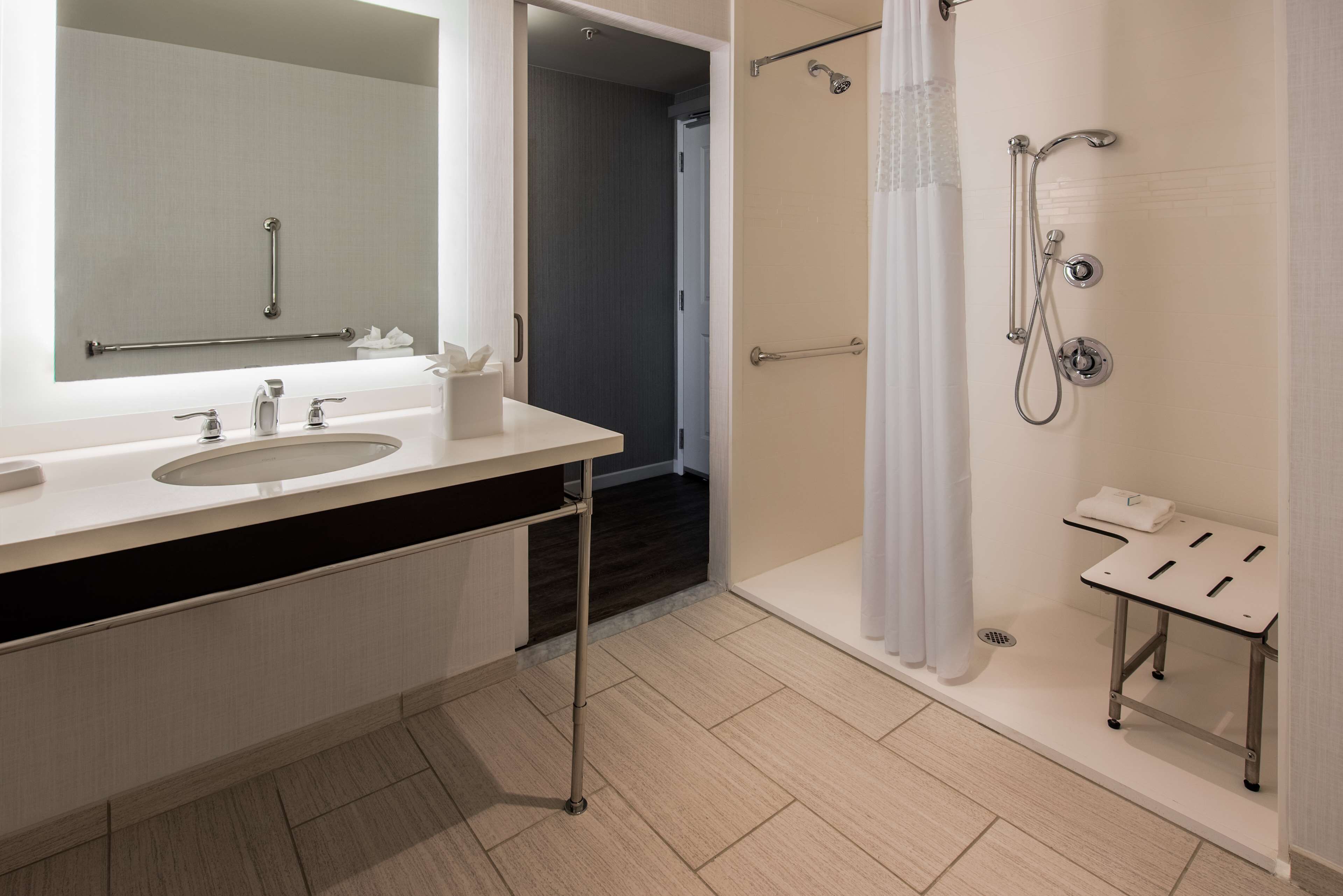 Une salle de bain moderne avec un meuble-lavabo à deux vasques, une douche équipée d’un pommeau de douche à main et une petite table sur laquelle est posée une serviette.