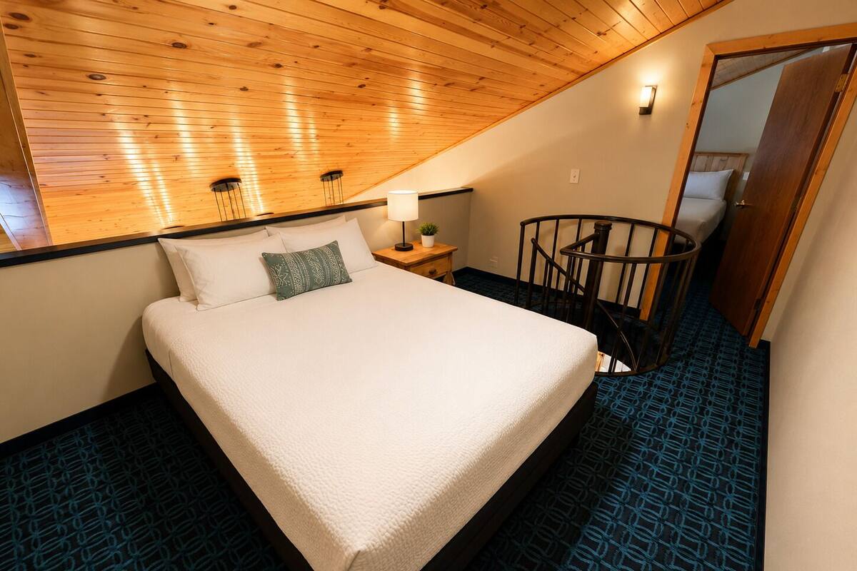 Une chambre avec un plafond en bois, un lit recouvert de linge de lit blanc, une table de chevet avec une lampe et un tapis bleu à motifs.