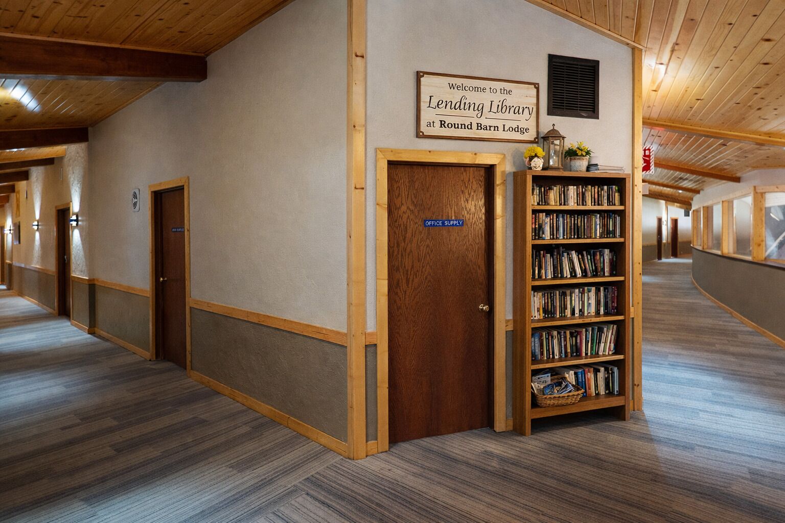 Un couloir avec une porte en bois portant l’inscription « Fournitures de bureau », une bibliothèque remplie de livres et un panneau indiquant : « Bienvenue à la bibliothèque de prêt du Round Barn Lodge. »