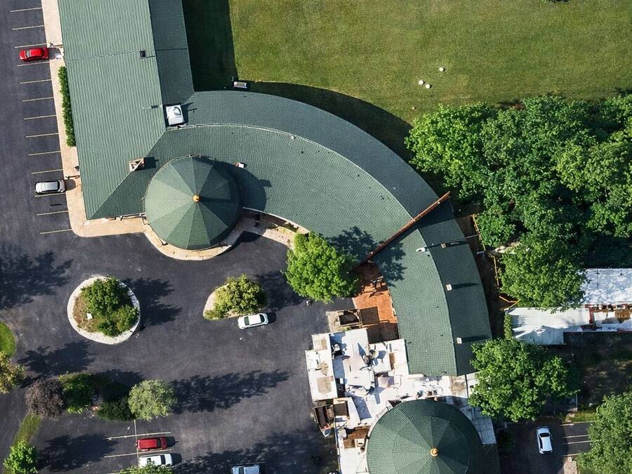 An aerial view of a building with a curved roof, multiple parking spaces, and surrounding greenery.