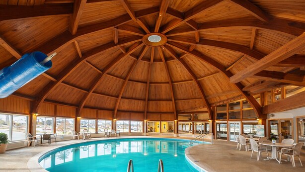 An indoor swimming pool with a wooden ceiling and surrounding seating areas. - The Round Barn Lodge (Spring Green)
