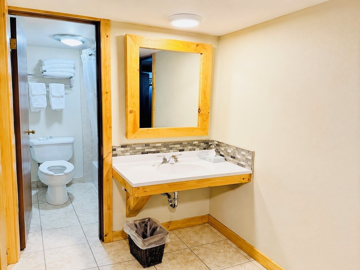 Un bagno con un lavandino bianco, un mobile vanitario in legno, uno specchio e un water visibile attraverso una porta aperta.