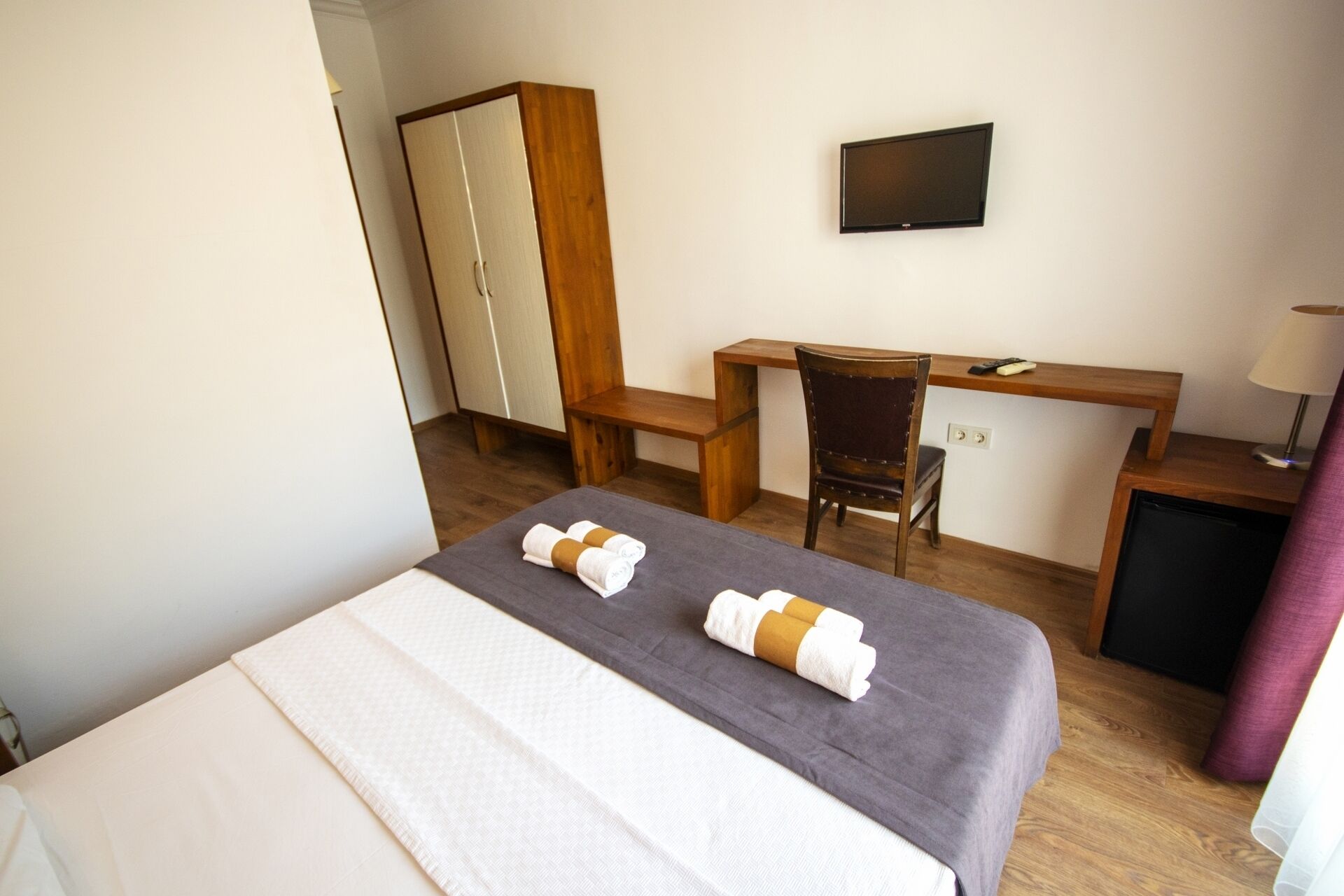 A hotel room with a bed, a desk, a chair, a wardrobe, a television, and a small table with a lamp.
