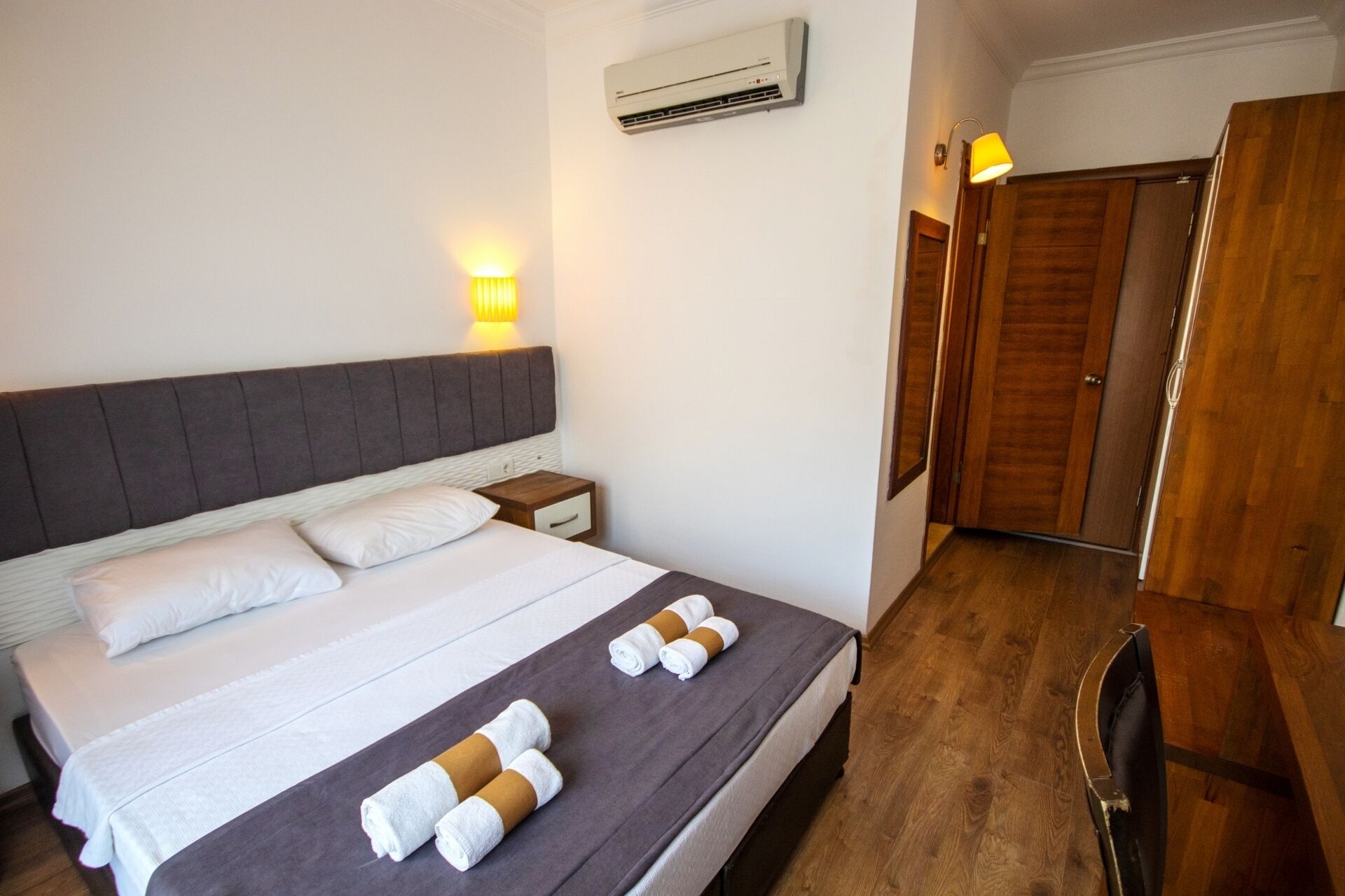 A hotel room with a bed, bedside table, wall-mounted air conditioner, and a wooden wardrobe.