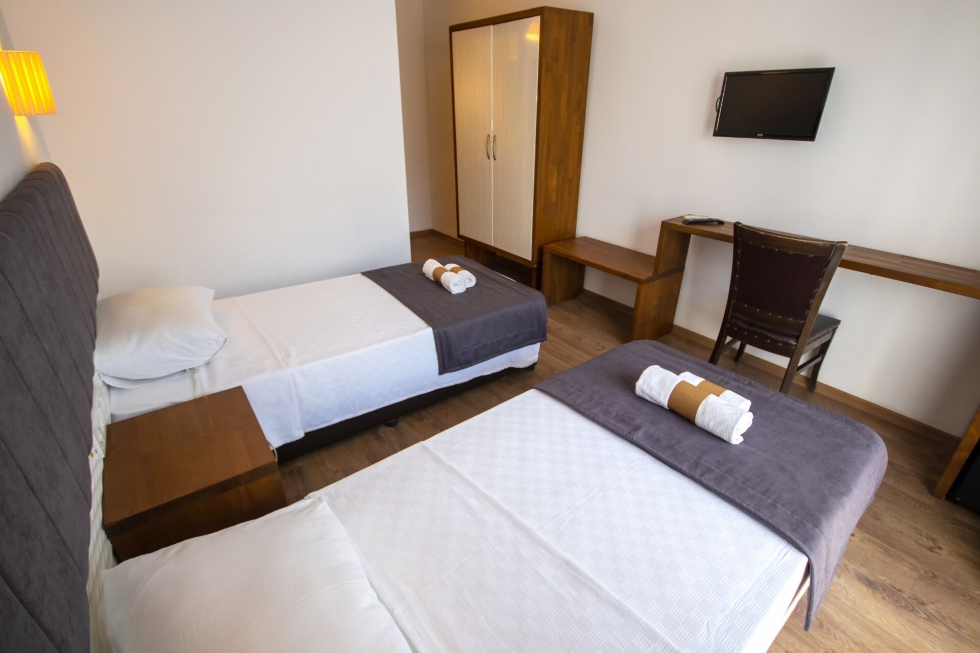 A hotel room with two beds, a wooden wardrobe, a desk, a chair, and a TV on the wall.