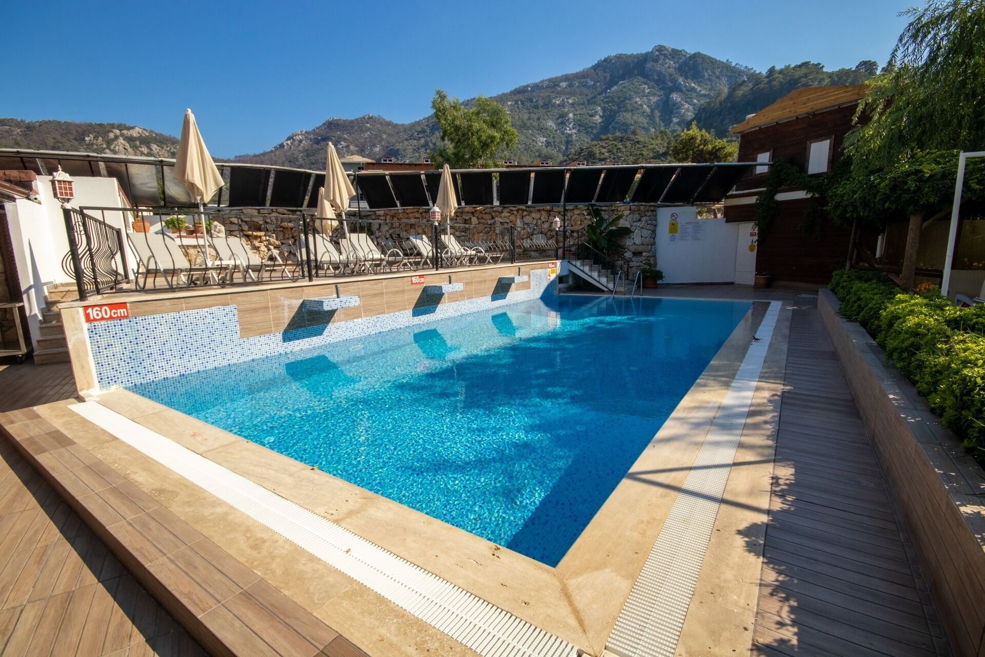 A rectangular outdoor swimming pool with clear blue water, surrounded by a wooden deck and a stone wall.