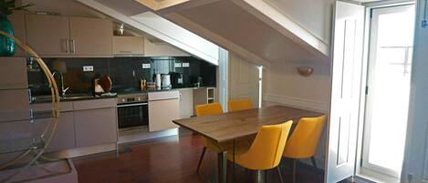 A modern kitchen with a dining area, featuring a wooden table and yellow chairs, and a large window providing natural light.
