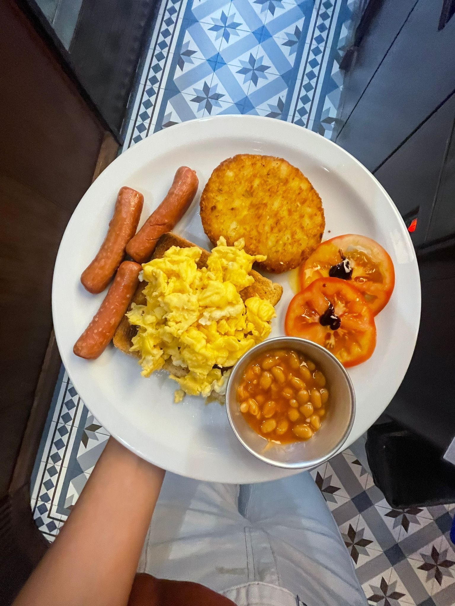 Un plato con huevos revueltos, salchichas, una hamburguesa, una rodaja de tomate y frijoles horneados.