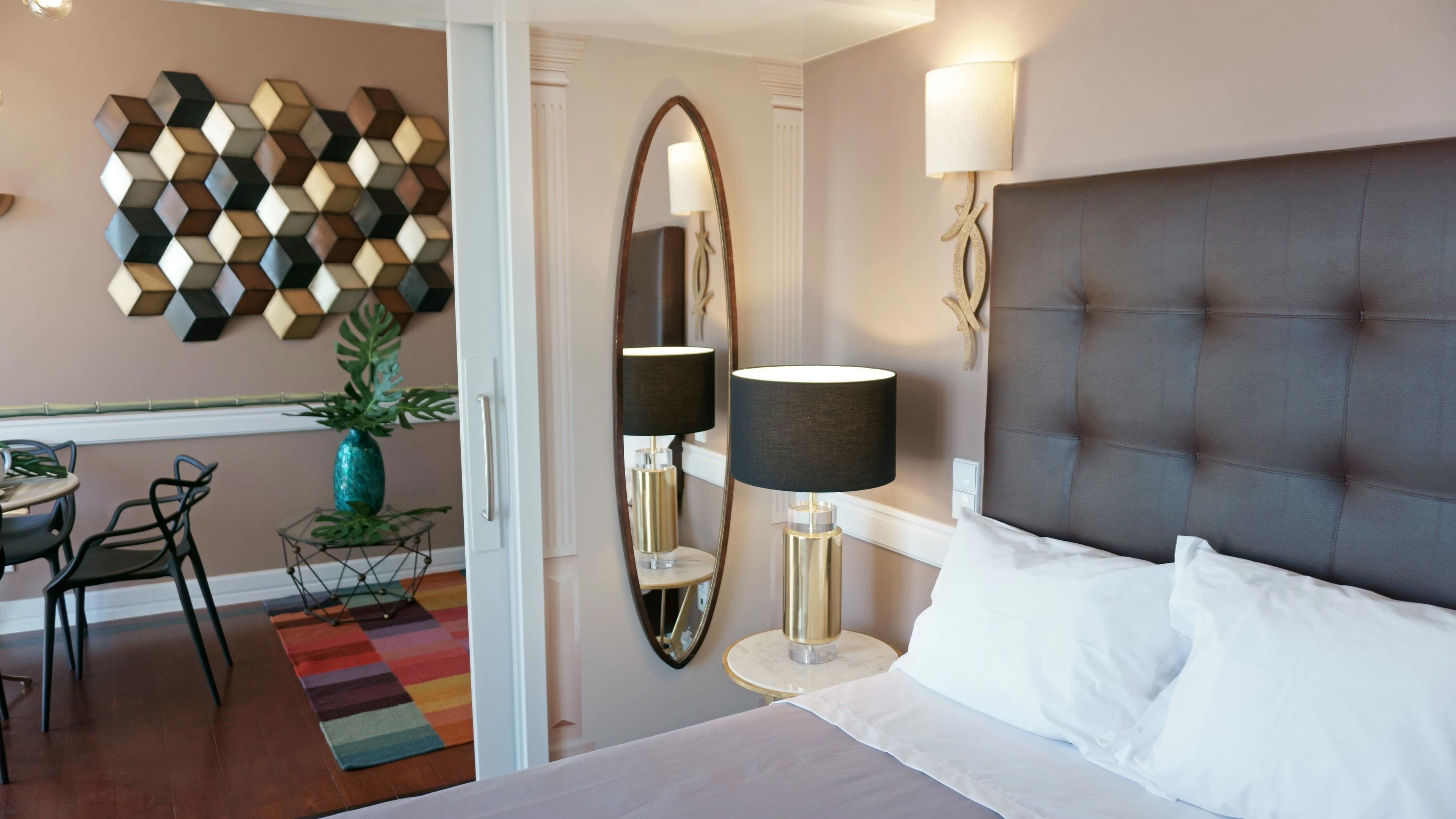 A hotel room with a bed, a large mirror, a lamp, a decorative wall piece, a small table with a vase, and a chair.