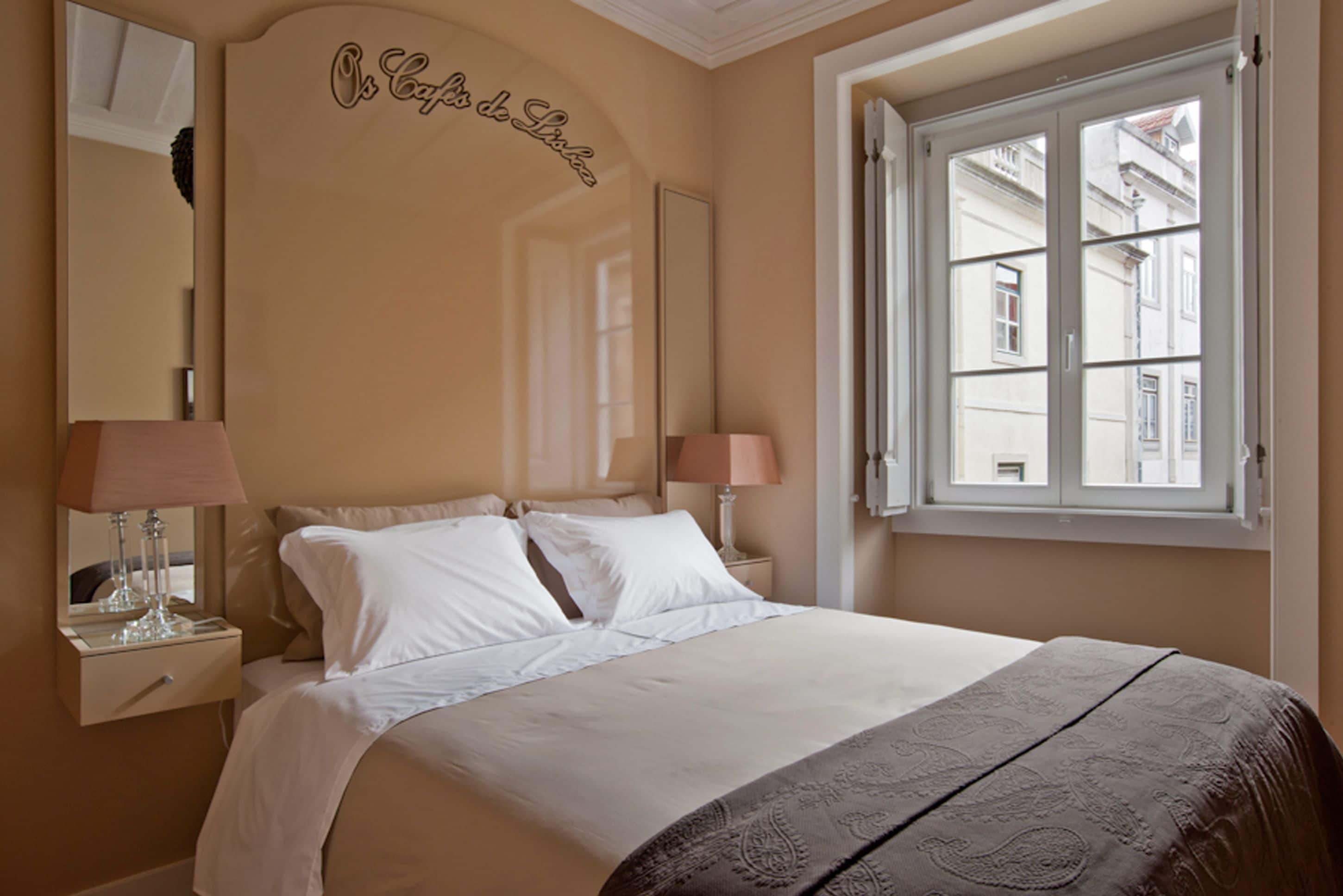 Ein Hotelzimmer mit einem großen Bett, zwei Nachttischlampen, einem Spiegel und einem Fenster mit Blick auf Gebäude.