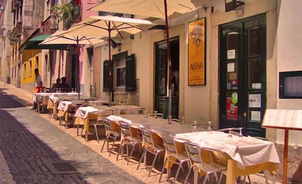 A row of outdoor tables and chairs with umbrellas, set against a building with a green door and a poster on the wall. - Lisbon Colours Bairro Alto Apartments (Lisbon)