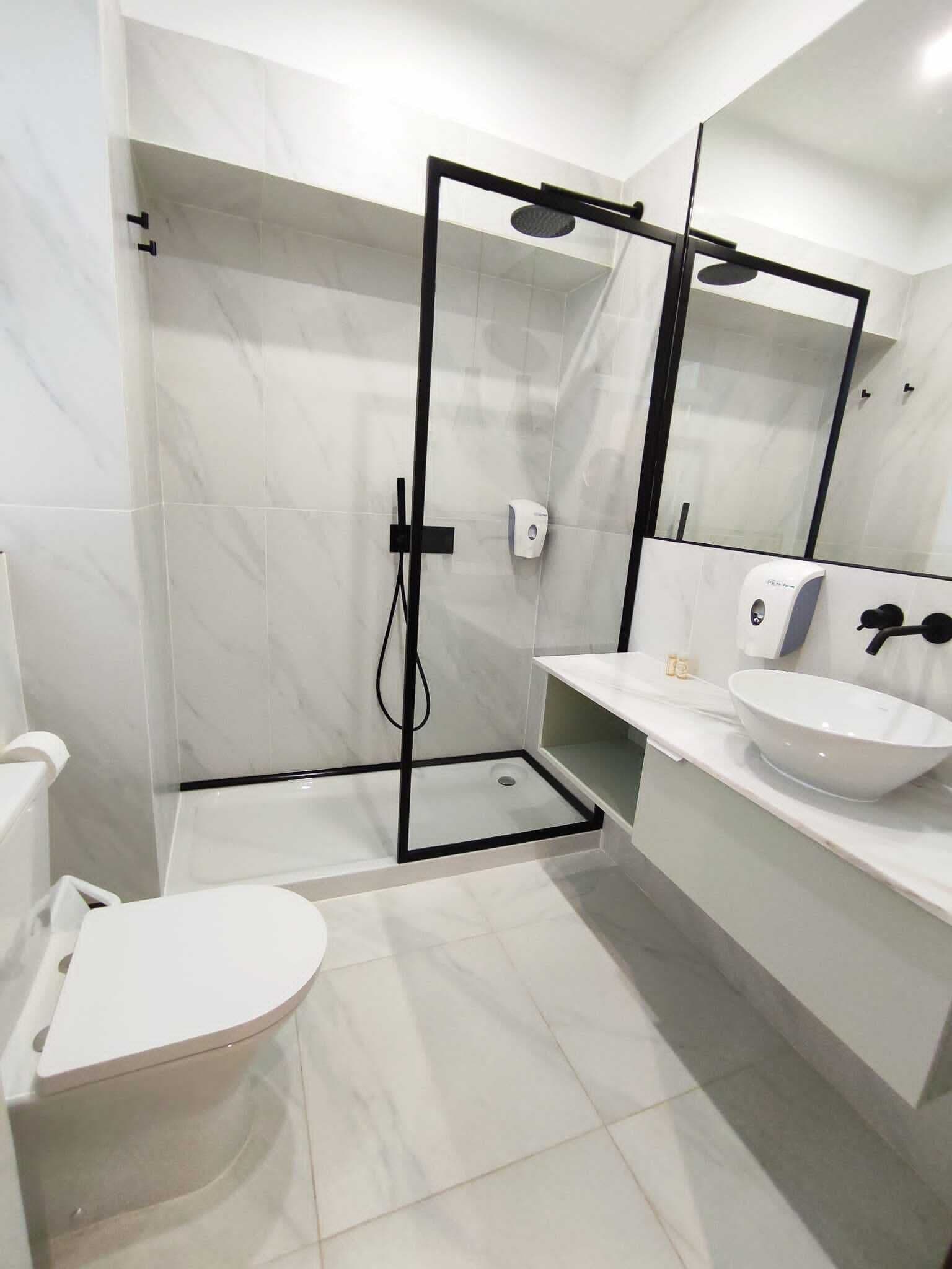 Un baño moderno con inodoro blanco, cabina de ducha con paredes de vidrio, lavamanos blanco y grifo negro, y un espejo grande.