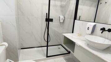 Un baño moderno con inodoro blanco, cabina de ducha con paredes de vidrio, lavamanos blanco y grifo negro, y un espejo grande.