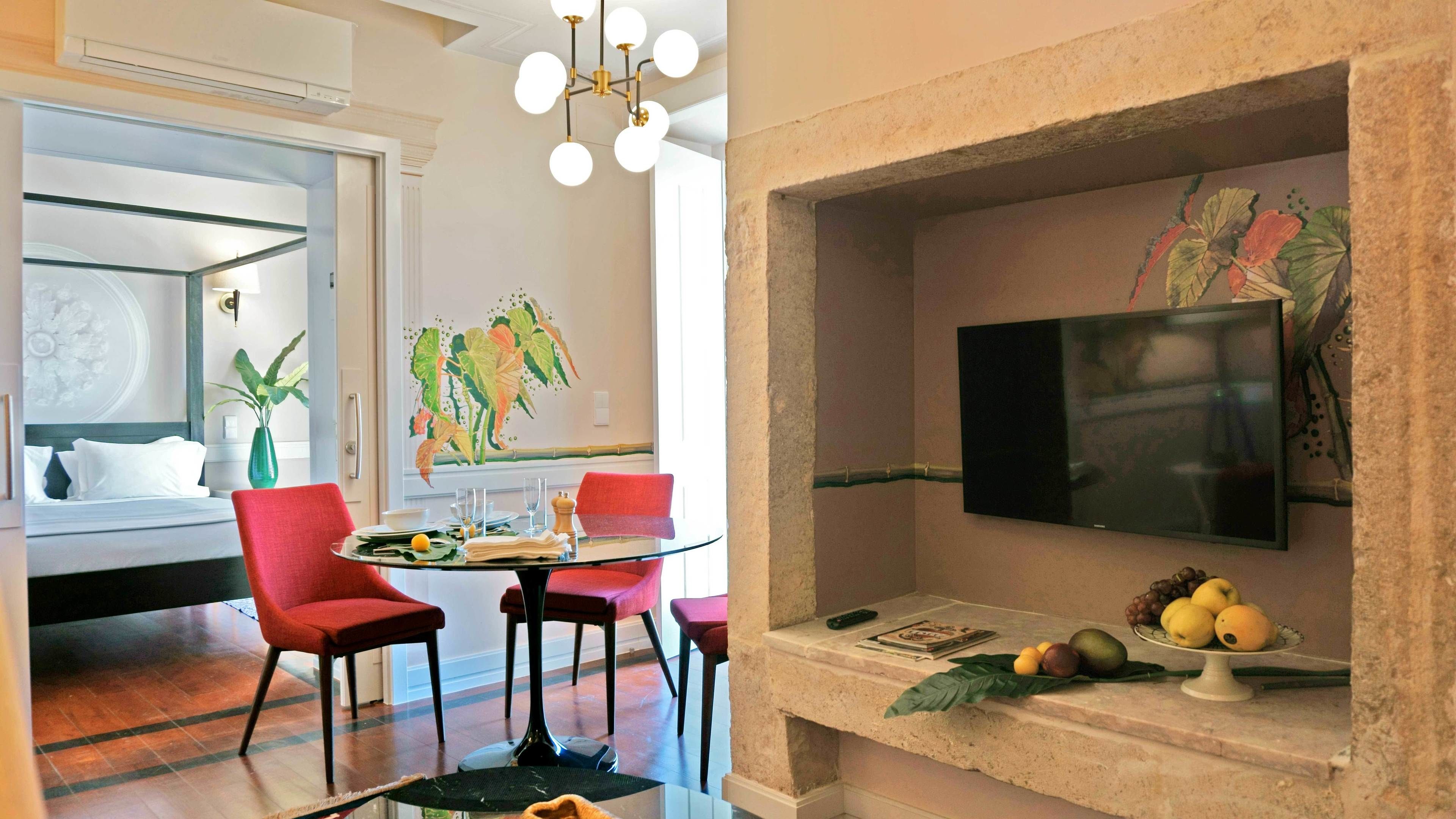 A modern hotel room with a dining area featuring a round glass table, red chairs, and a flat-screen TV mounted on a stone wall.