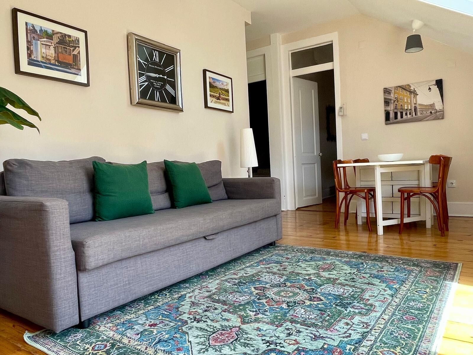 A living room with a grey sofa, green cushions, a patterned rug, a dining table with chairs, and framed pictures on the wall.
