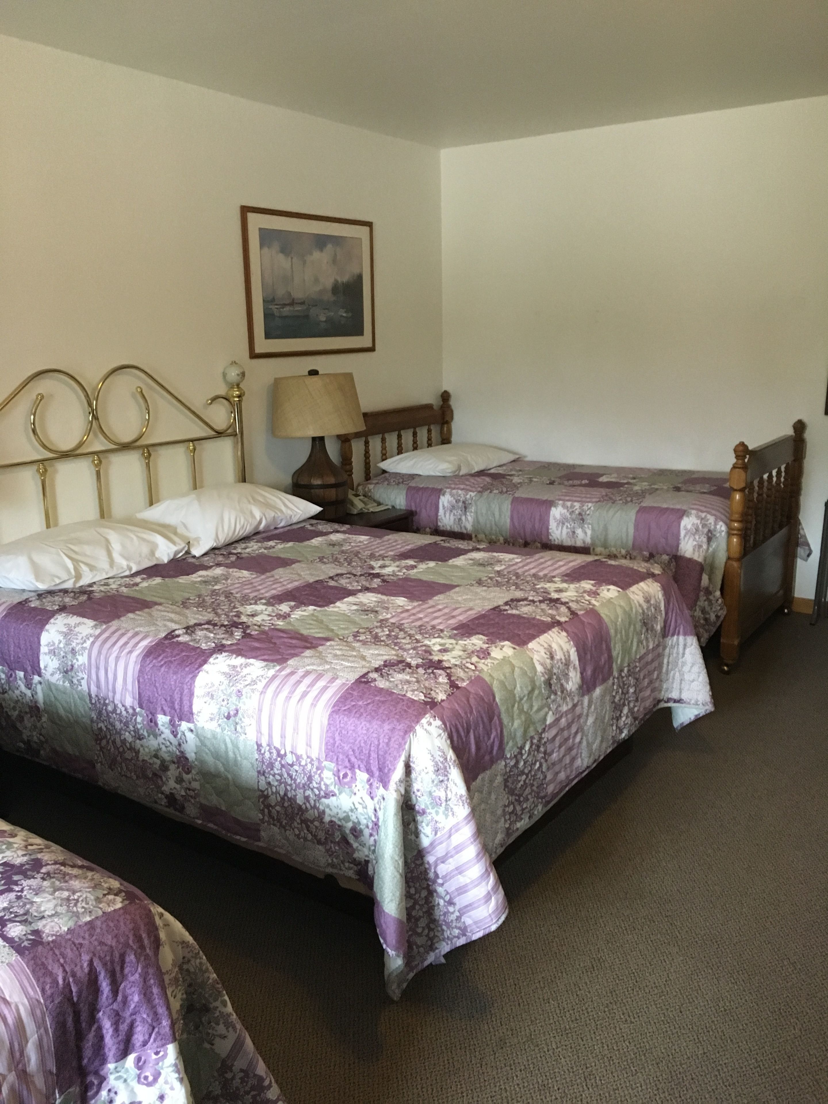 A room with two single beds, each with a wooden headboard and a floral-patterned bedspread. There is a nightstand with a lamp between the beds, and a framed picture on the wall above the headboard.