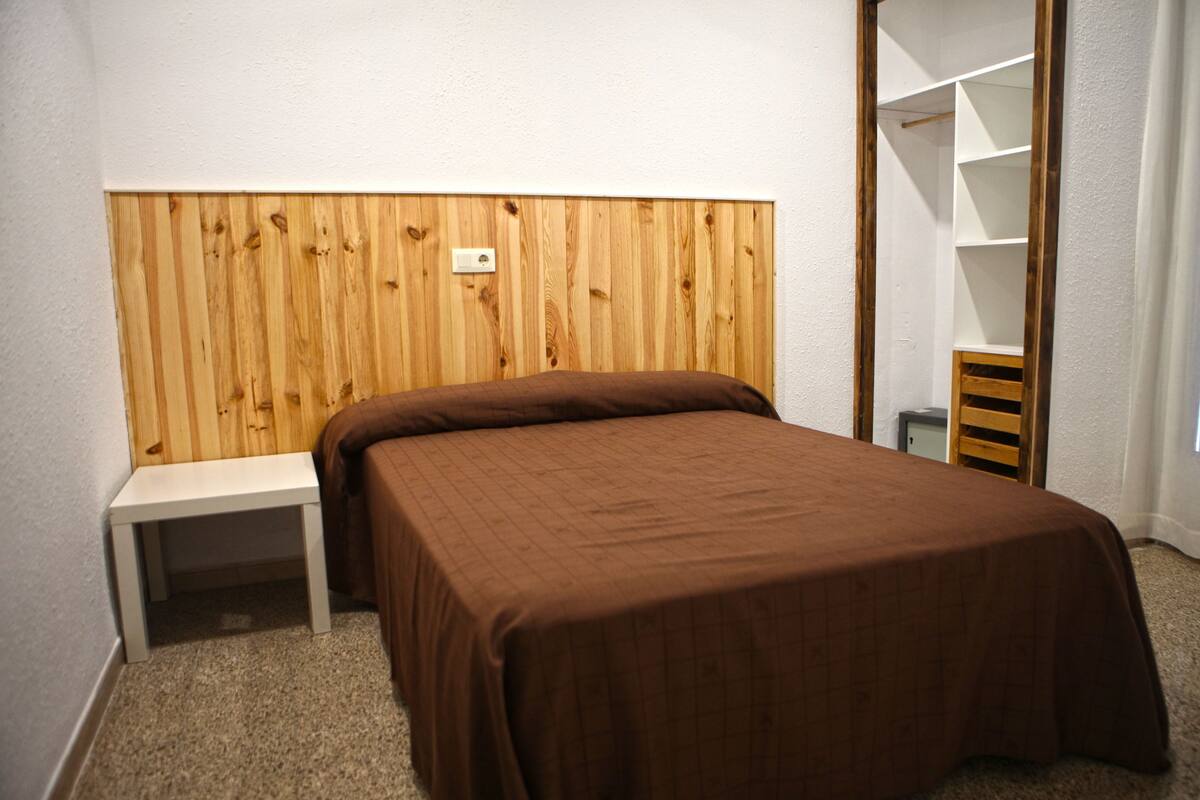 A bedroom with a wooden headboard, a single bed with a brown bedspread, a small white nightstand, and a built-in wardrobe with open shelves.