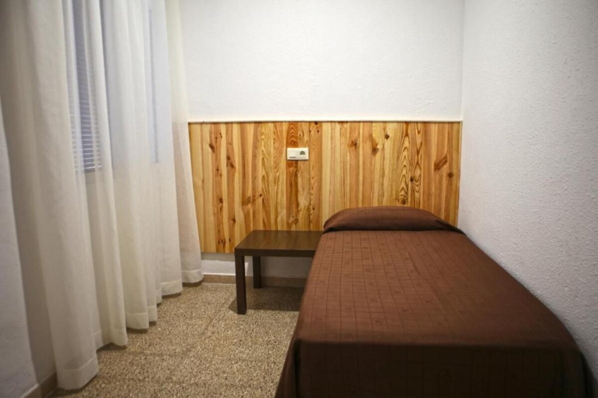 Una habitación pequeña con cama individual, cabecera de madera, mesita de noche y ventana con cortinas blancas.