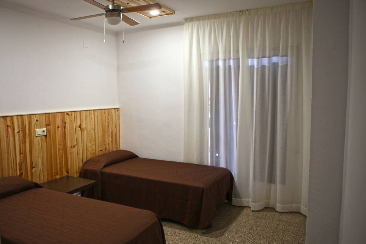 A room with two single beds, a wooden panel wall, a ceiling fan, and a window with sheer curtains.