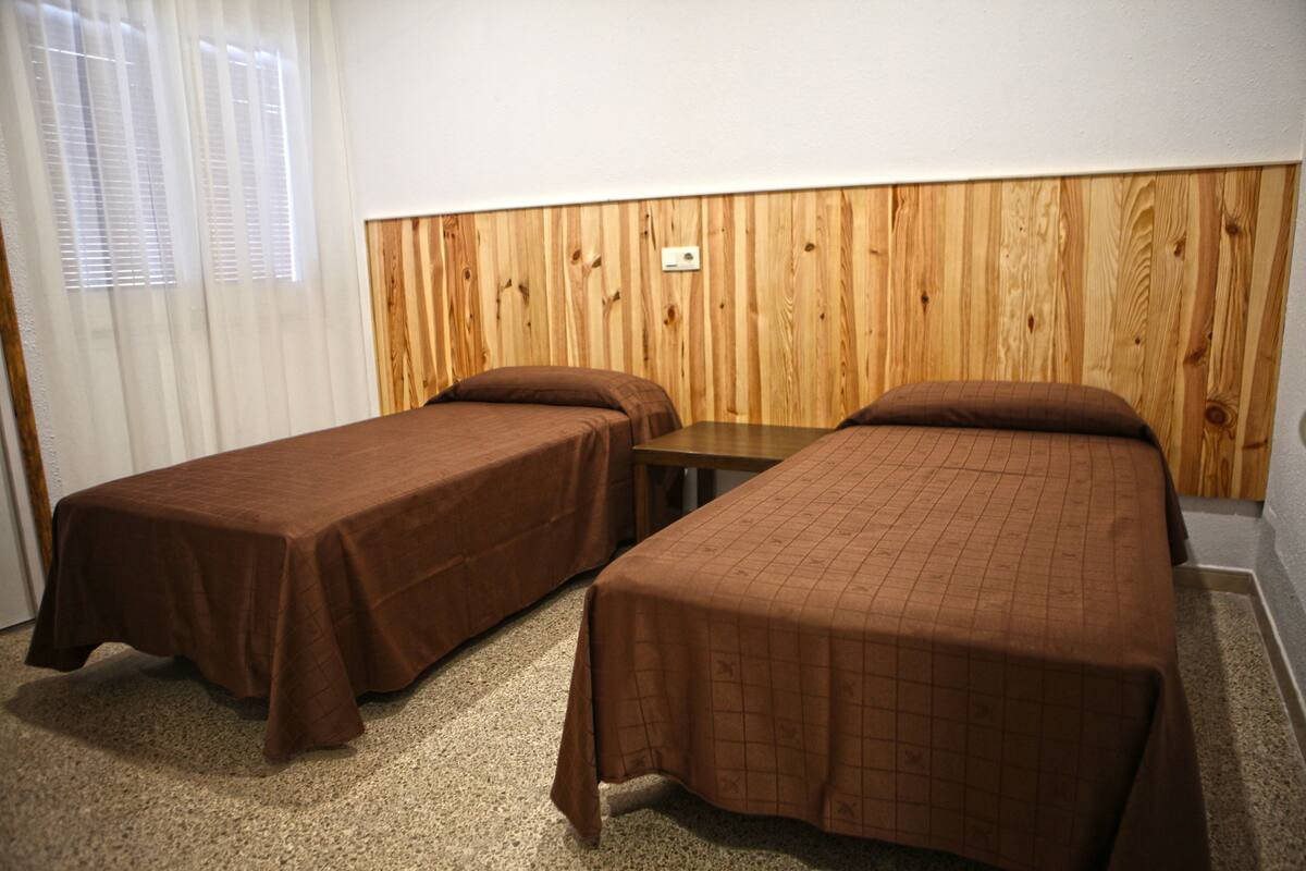 A room with two single beds, a wooden headboard, a small table, and a window with blinds.