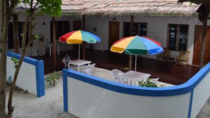 A covered outdoor area with white plastic chairs and tables, two colorful umbrellas, and a thatched roof.