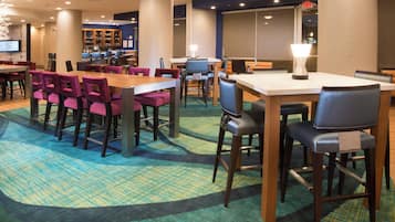 Un hall d'hôtel doté de plusieurs espaces aménagés avec des sièges, notamment une longue table avec des chaises violettes et un espace séparé comprenant un bar et des tables individuelles.