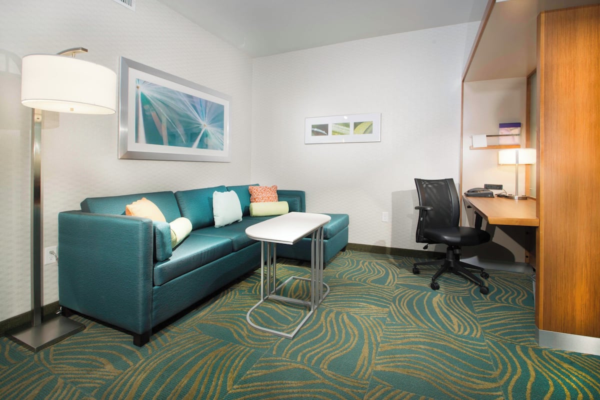 A hotel room with a teal sofa, a small white table, a black office chair, a desk with a lamp, and a patterned carpet.