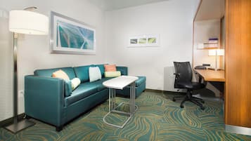 Une chambre d’hôtel avec un canapé de couleur turquoise, une petite table blanche, un fauteuil de bureau noir, un bureau avec une lampe et une moquette à motifs.