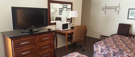 A hotel room with a flat-screen TV, a microwave, a desk with a chair, two beds with patterned bedspreads, a mirror, and a wall-mounted shelf.