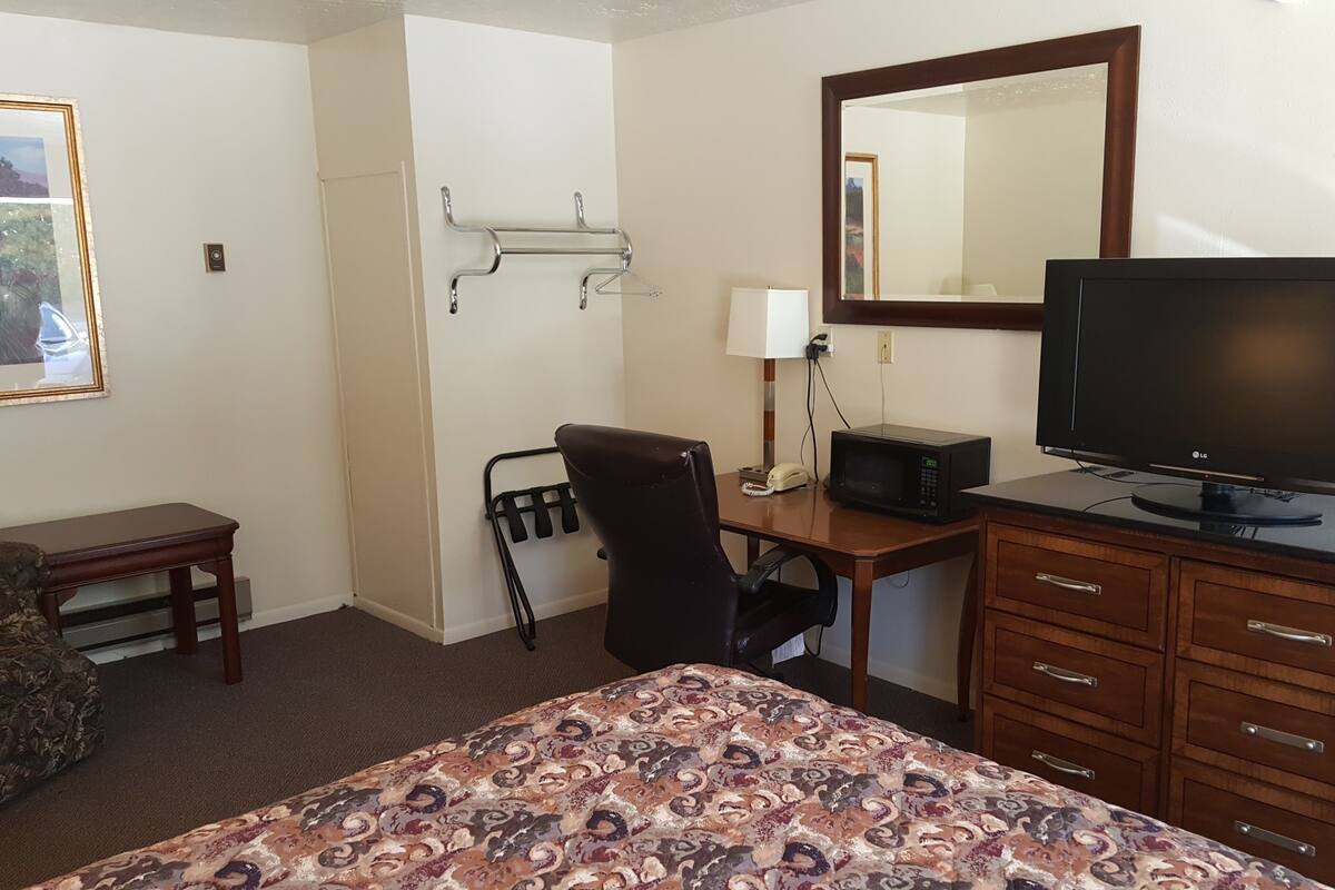 Ett hotellrum med en säng, ett skrivbord med en dator, en tv, en stol, en telefon och en klädställning.