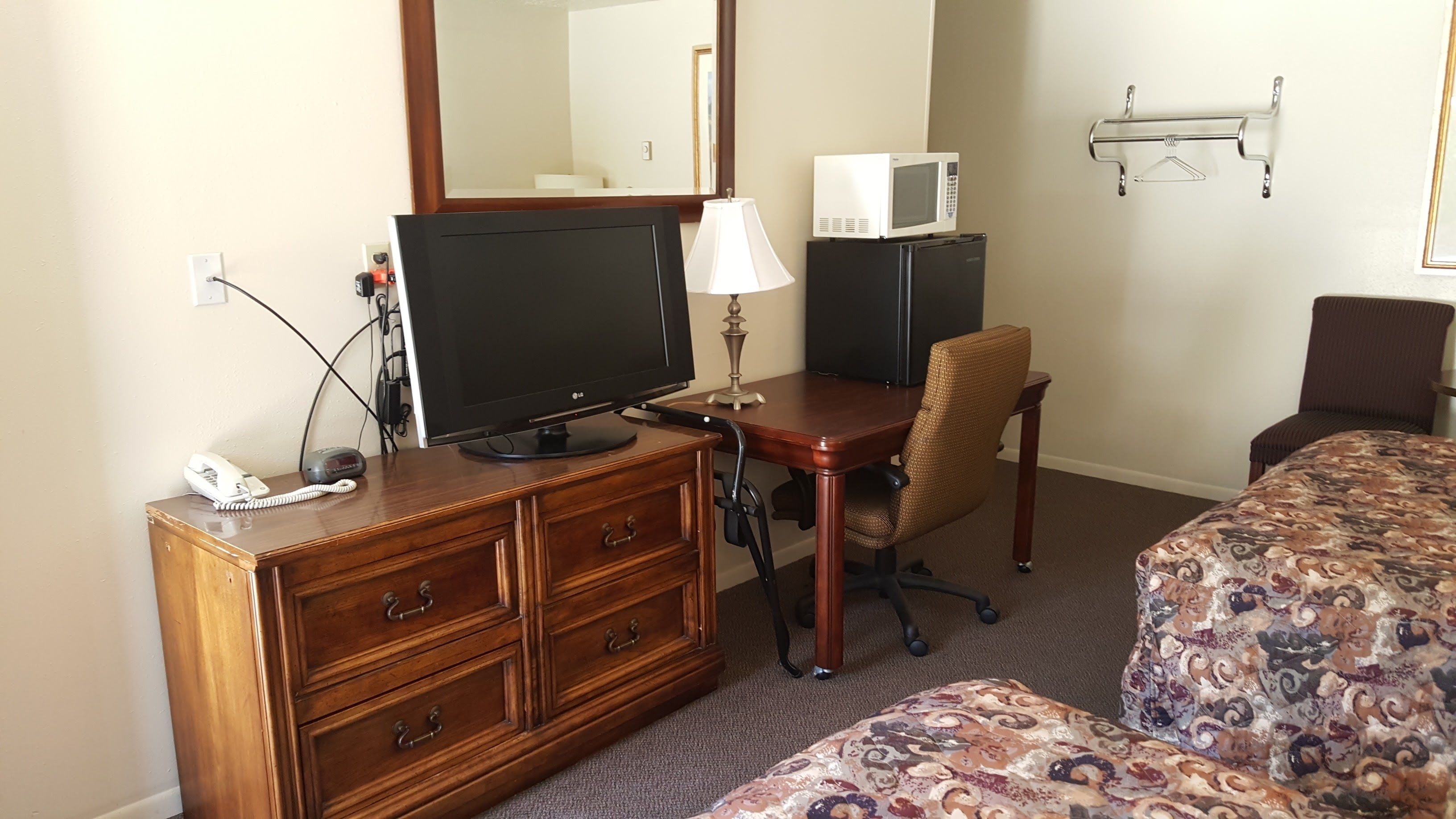 A hotel room with a wooden dresser, a desk with a television, a microwave, a chair, and a bed with patterned bedding.