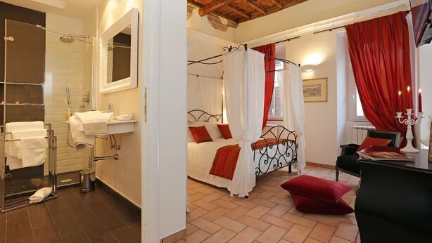 A bedroom with a bed, a nightstand, a chair, a window with red curtains, a shower, and a bathroom with a sink and mirror. - Madonna de’ Monti Suites  (Rome)