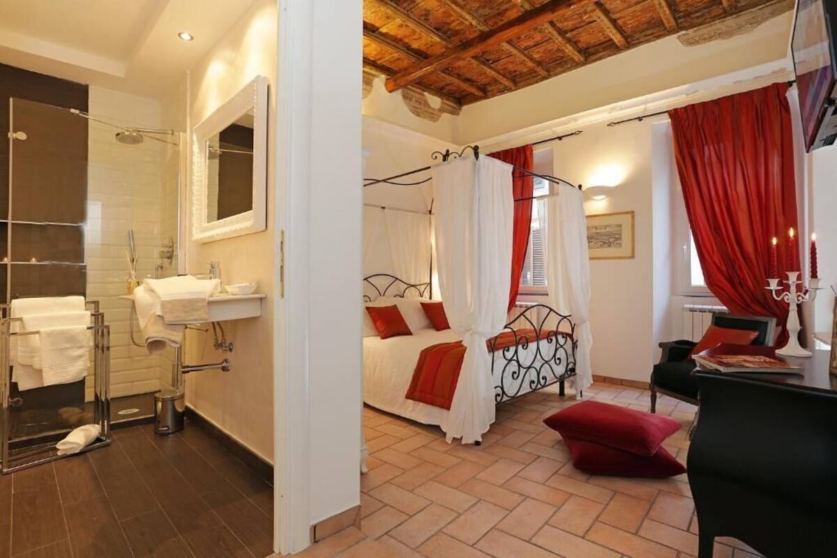 A bedroom with a bed, a nightstand, a chair, a window with red curtains, a shower, and a bathroom with a sink and mirror.