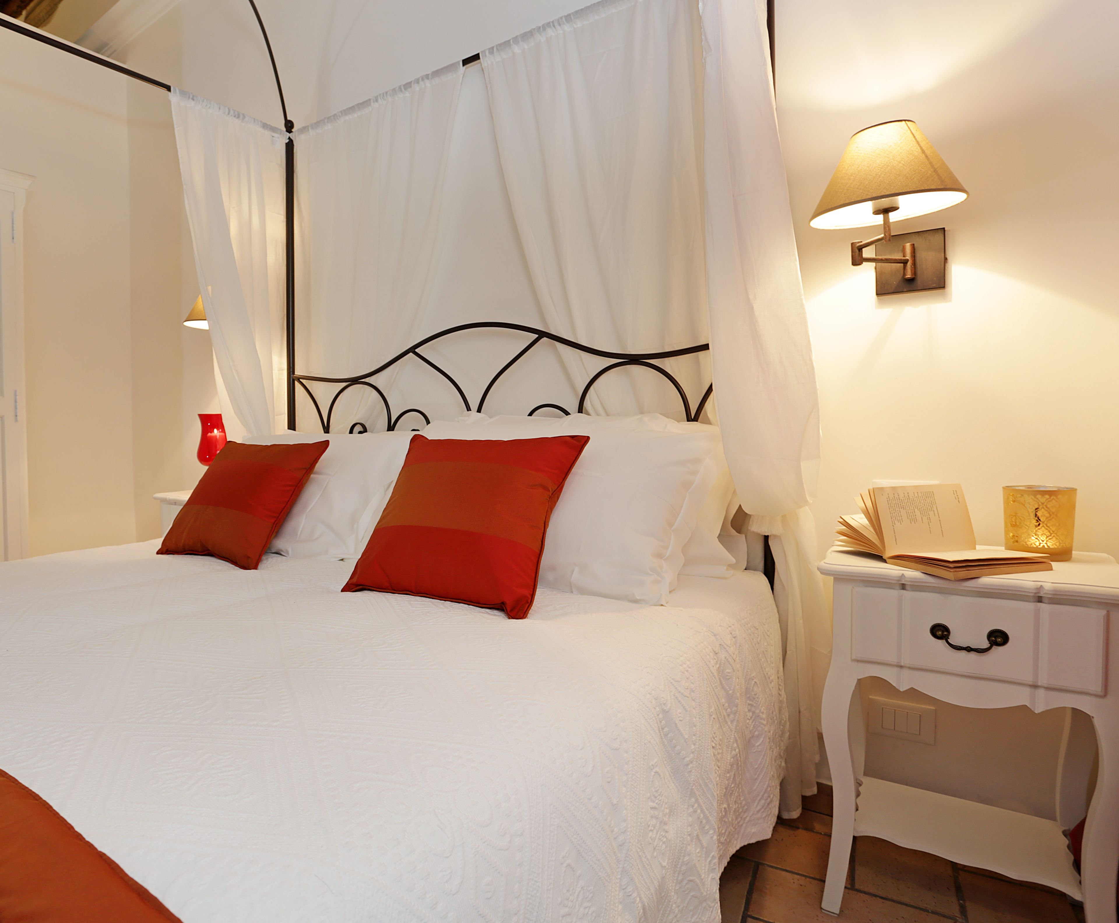 A neatly made bed with white linens and two red pillows, a bedside table with a lamp, and a small chair.