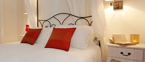 A neatly made bed with white linens and two red pillows, a bedside table with a lamp, and a small chair.