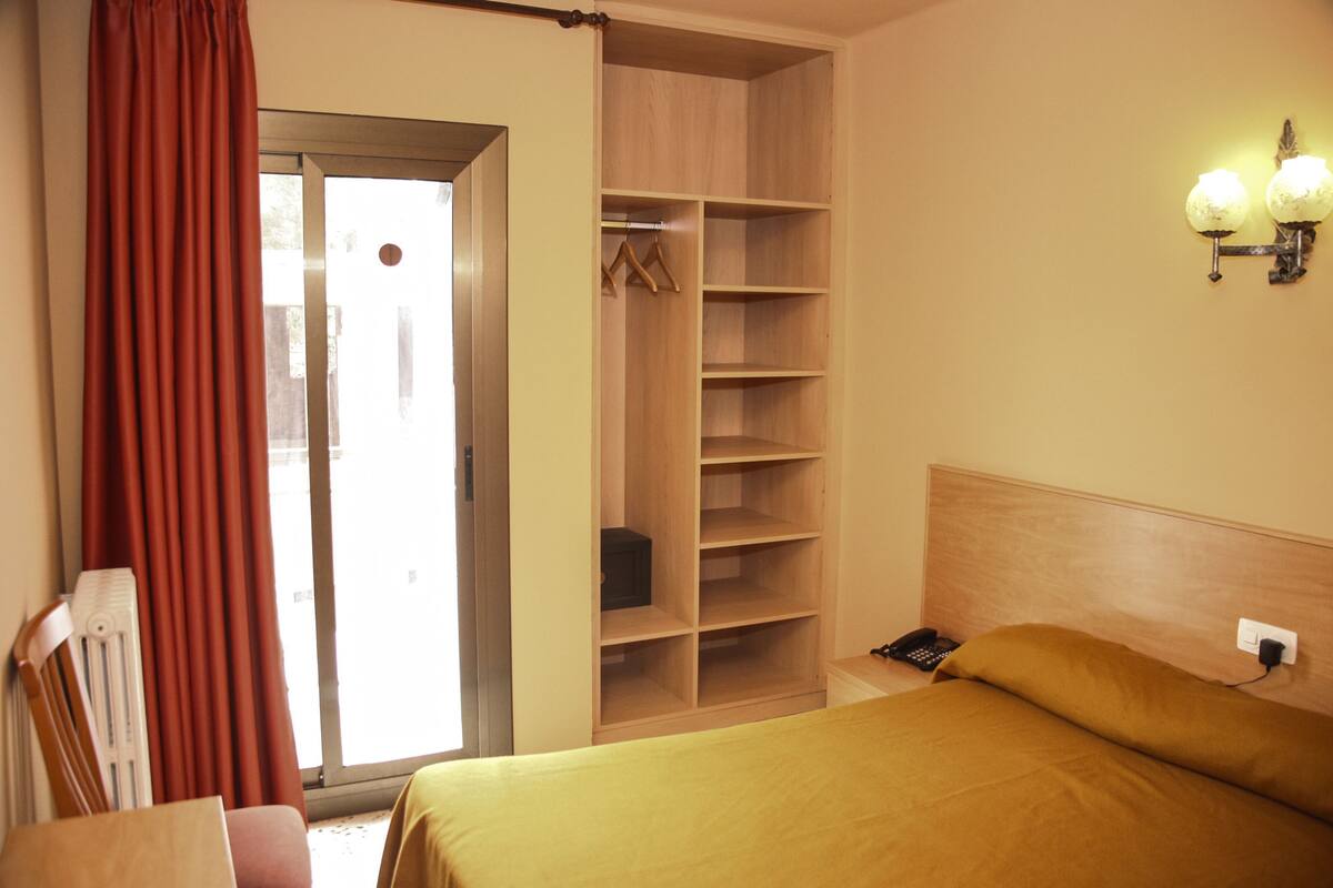 A hotel room with a bed, a wooden wardrobe, a desk, and a chair.
