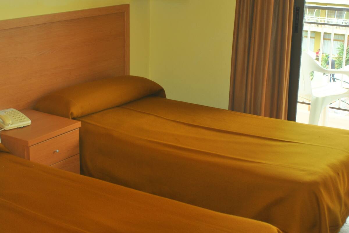 A hotel room with two beds, a wooden headboard, a bedside table with a telephone, and a view of the outside balcony.