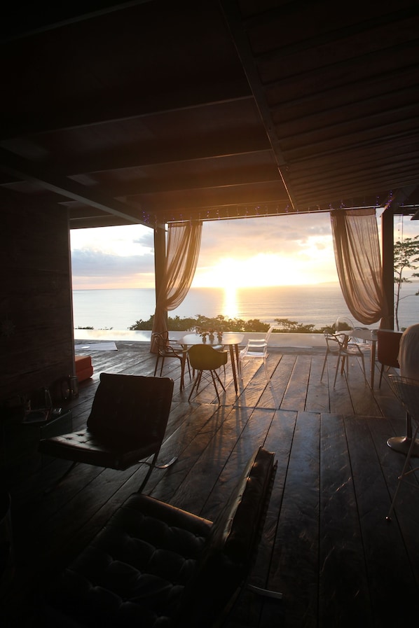 Une terrasse en bois offrant une vue sur le coucher de soleil sur l’océan, avec une table et des chaises.