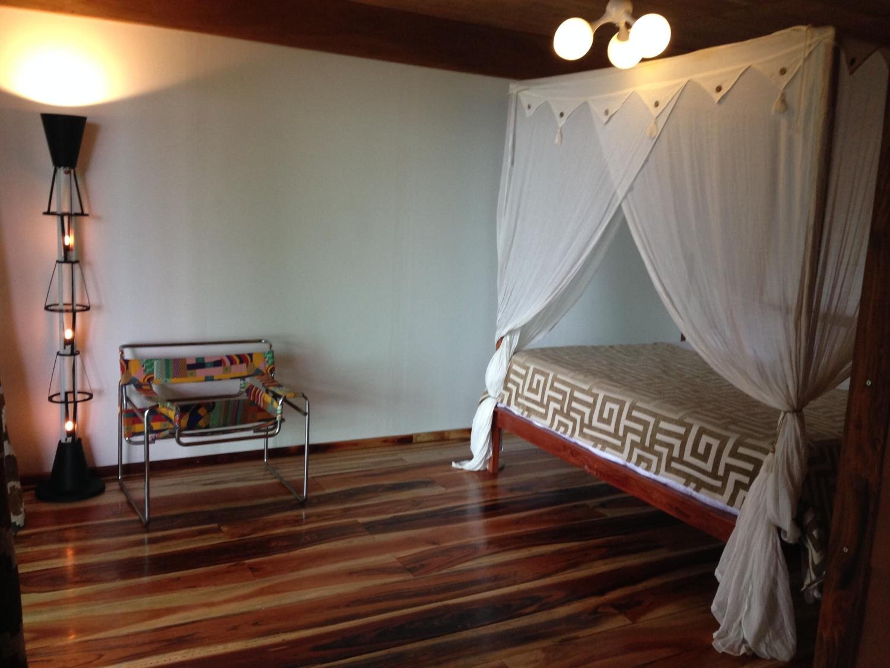 A room with a wooden floor, a bed with a white canopy, a colorful bench, and a floor lamp.