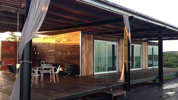 Een overdekt buitengebied met een houten terras, een tafel met stoelen en uitzicht op een natuurlandschap door grote glazen deuren.