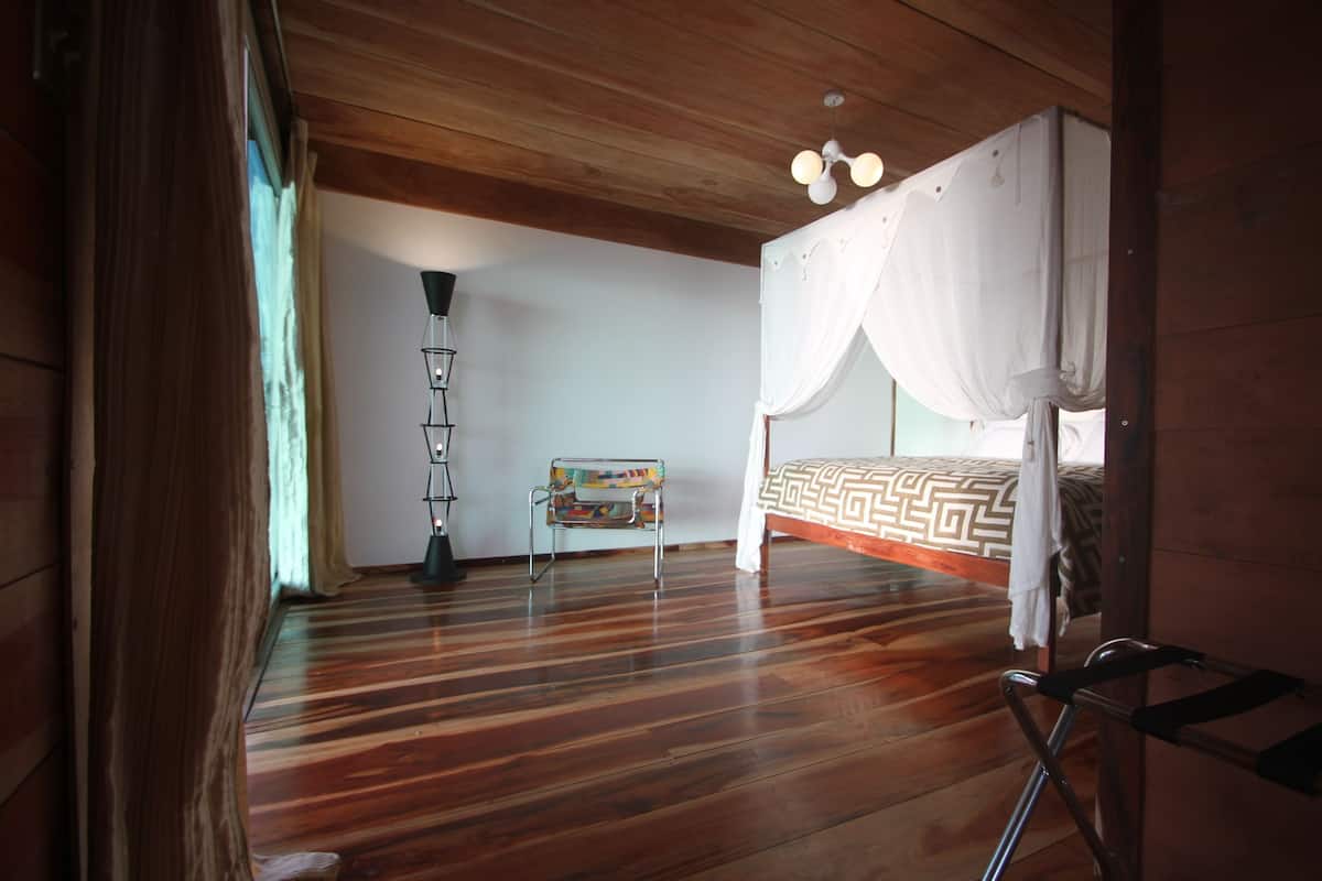 Um quarto com uma cama, uma cadeira, um abajur e piso de madeira.